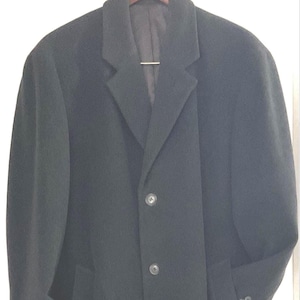 May include: A solid black overcoat featuring a notched lapel, a two-button front closure, and long sleeves with buttoned cuffs. The coat is constructed with a structured collar and a smooth, dark fabric finish, hanging straight from the shoulders.