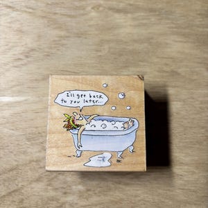 May include: A wooden stamp featuring a cartoon illustration of a person relaxing in a clawfoot bathtub filled with bubbles. The text "I'll get back to you later..." is above the tub. The stamp is on a wooden surface.