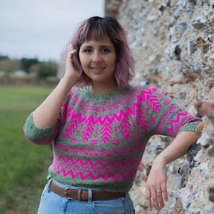 Könnte beinhalten: Eine Frau trägt einen grün-pinken Pullover mit geometrischem Muster, blaue Jeans und schwarze Turnschuhe. Sie steht neben einer Steinmauer.