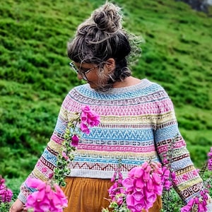 Pode incluir: Uma pessoa vestindo um suéter listrado colorido e calças marrons está em um campo de flores de dedaleira rosa. As flores estão em flor e a pessoa está olhando para elas.