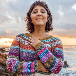 Może przedstawiać: Kolorowy sweter we wzory w odcieniach pomarańczu, różu, błękitu i zieleni. Jest zestawiony z długą, beżową spódnicą z detalami guzików. Tło przedstawia scenę na plaży z oceanem i zachodem słońca.