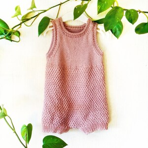 May include: A pink knitted baby onesie with a sleeveless design, hanging on a white hanger against a white wall with a green plant in the background.