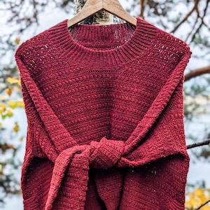 Może przedstawiać: Bordowy sweter bez rękawów z okrągłym dekoltem i wiązanym detalem w talii. Sweter wykonany jest z teksturowanej dzianiny i wisi na drewnianym wieszaku. Tło przedstawia rozmyty krajobraz zewnętrzny.