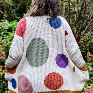 May include: Cream-colored knit cardigan with large, colorful polka dots in red, blue, green, purple, and orange. The cardigan has a mustard yellow trim and is worn outdoors.