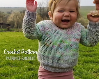 May include: A toddler wearing a colorful crochet sweater and pink leggings. The sweater has a speckled pattern with shades of green, blue, and pink. The child is smiling and holding onto an adult's hand. The background is a grassy field.