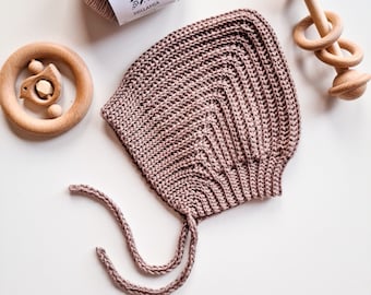 May include: A dusty rose crocheted baby bonnet with a tie string. The bonnet is laying on a white surface next to a ball of yarn and a wooden baby rattle.