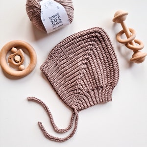 Könnte beinhalten: Eine staubige rosafarbene gehäkelte Babyhaube mit einer Bindekordel. Die Haube liegt auf einer weißen Oberfläche neben einer Garnrolle und einer Holzrassel.