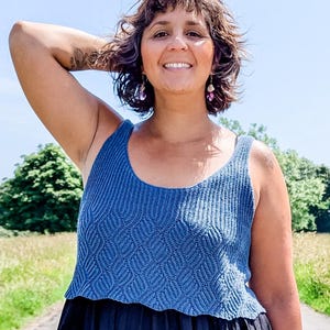 Puede incluir: Una persona con un top azul tejido y una falda larga negra y fluida de pie en una carretera. El top tiene un patrón texturizado, y la falda llega hasta los tobillos. Sandalias marrones completan el atuendo, con campos verdes y cielo azul de fondo.