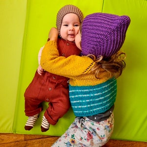 Pu&ograve; includere: Due bambini con cappelli di lana. Il bambino a sinistra indossa un cappello marrone e un vestito rosso e marrone. Il bambino a destra indossa un cappello viola e un maglione a righe giallo, blu e bianco. I bambini si abbracciano e sorridono.