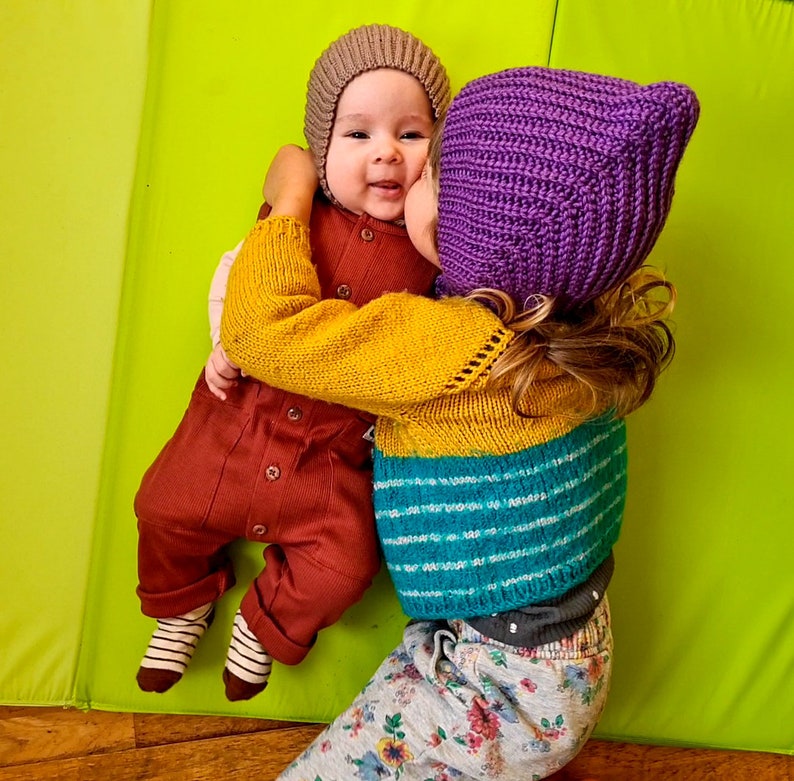 Pu&ograve; includere: Due bambini con cappelli di lana. Il bambino a sinistra indossa un cappello marrone e un vestito rosso e marrone. Il bambino a destra indossa un cappello viola e un maglione a righe giallo, blu e bianco. I bambini si abbracciano e sorridono.