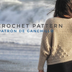 Puede incluir: Una mujer lleva un suéter de crochet beige con un patrón texturizado. El suéter tiene un corte holgado y mangas largas. El texto "CROCHET PATTERN PATRÓN DE GANCHILLO" es visible en la imagen.
