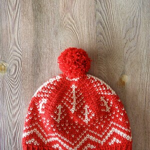 Puede incluir: Gorro de punto rojo con un patr&oacute;n geom&eacute;trico blanco y un pomp&oacute;n rojo en la parte superior.