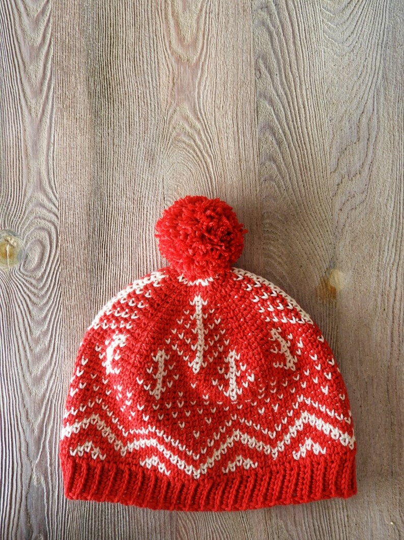 Puede incluir: Gorro de punto rojo con un patr&oacute;n geom&eacute;trico blanco y un pomp&oacute;n rojo en la parte superior.