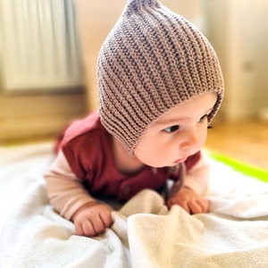 Pu&ograve; includere: Un bambino con un berretto di lana marrone &egrave; sdraiato su una coperta bianca. Il bambino sta guardando di lato e ha le mani sulla coperta.