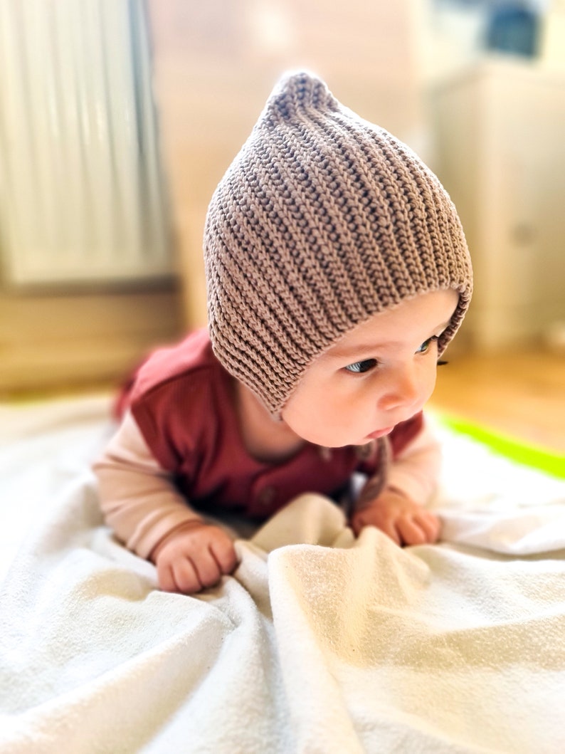 Pu&ograve; includere: Un bambino con un berretto di lana marrone &egrave; sdraiato su una coperta bianca. Il bambino sta guardando di lato e ha le mani sulla coperta.