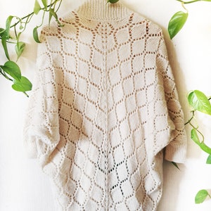 Op de afbeelding: Een crèmekleurige gehaakte cardigan met een ruitpatroon. De cardigan heeft een hoge hals en lange mouwen.