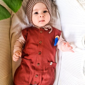 Pu&ograve; includere: Un bambino che indossa un berretto di lana marrone e un body a costine color ruggine con bottoni sul davanti. Il bambino &egrave; sdraiato in una culla bianca.