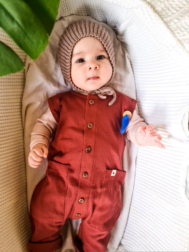 Pu&ograve; includere: Un bambino che indossa un berretto di lana marrone e un body a costine color ruggine con bottoni sul davanti. Il bambino &egrave; sdraiato in una culla bianca.