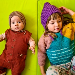 Pu&ograve; includere: Due bambini che indossano cappelli di lana. Il bambino a sinistra indossa un cappello marrone e una tuta marrone. Il bambino a destra indossa un cappello viola e un maglione multicolore con un motivo floreale.