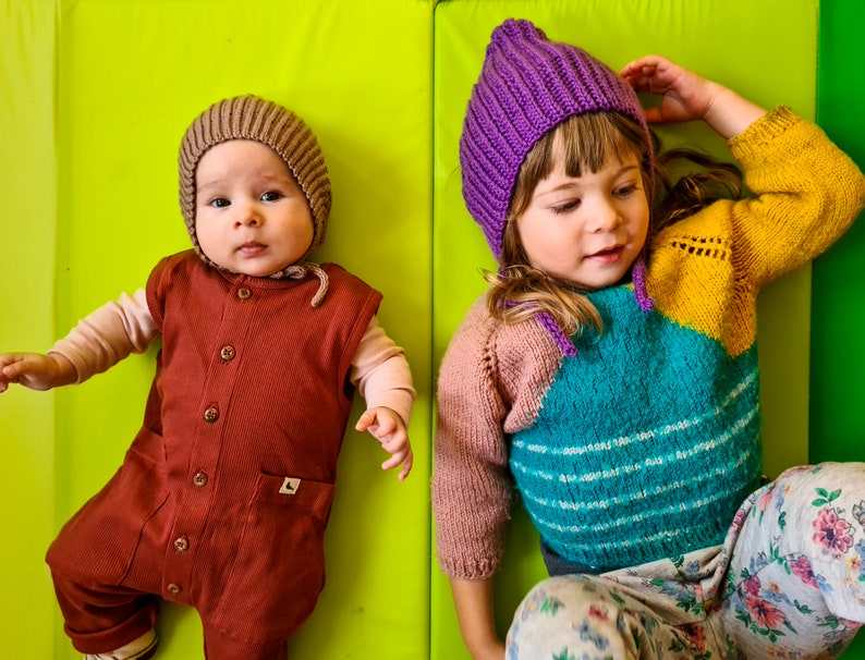 Pu&ograve; includere: Due bambini che indossano cappelli di lana. Il bambino a sinistra indossa un cappello marrone e una tuta marrone. Il bambino a destra indossa un cappello viola e un maglione multicolore con un motivo floreale.