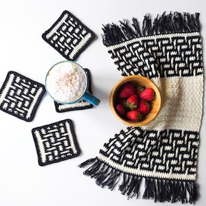 May include: Four black and white crocheted coasters with a geometric pattern, a blue mug with a cappuccino, a bowl of strawberries, and a black and white crocheted runner with fringe.