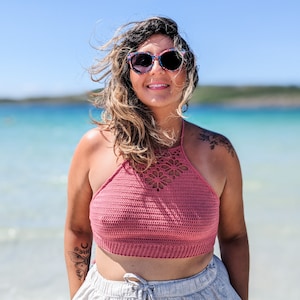 Könnte beinhalten: Eine Frau trägt ein pinkfarbenes gehäkeltes Halstuch und eine graue Leinenhose. Sie steht an einem Strand mit blauem Wasser im Hintergrund. Sie trägt eine Sonnenbrille und ihre Haare wehen im Wind.