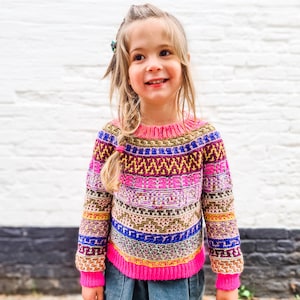 Puede incluir: Una niña pequeña con un suéter de rayas de colores con un patrón rosa, amarillo, azul, verde y morado. También lleva una falda azul oscuro y tiene el pelo rubio.