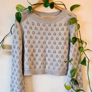 May include: A light grey knitted jumper with a delicate floral pattern. The jumper has a round neckline and long sleeves. It is hanging on a wooden hanger.