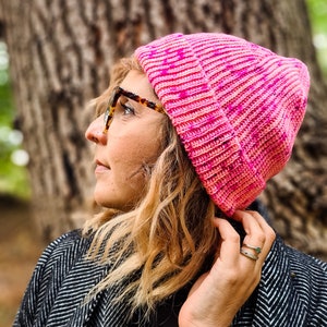 Könnte beinhalten: Eine Frau trägt eine rosafarbene Strickmütze mit einem weißen und rosafarbenen Farbmuster. Sie trägt eine Brille und einen schwarz-weiß gemusterten Mantel.