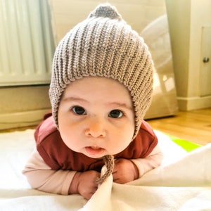 Pu&ograve; includere: Un bambino che indossa un berretto di lana marrone chiaro con la punta a punta. Il bambino &egrave; sdraiato su una coperta bianca e guarda la telecamera.