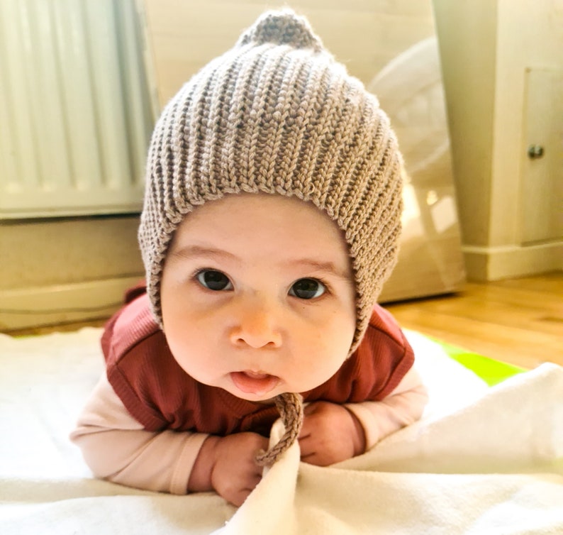 Pu&ograve; includere: Un bambino che indossa un berretto di lana marrone chiaro con la punta a punta. Il bambino &egrave; sdraiato su una coperta bianca e guarda la telecamera.