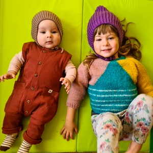 Pu&ograve; includere: Due bambini con cappelli di lana. Il bambino a sinistra indossa un cappello di lana marrone e una tuta marrone. Il bambino a destra indossa un cappello di lana viola e un maglione a righe colorate con pantaloni a fiori.