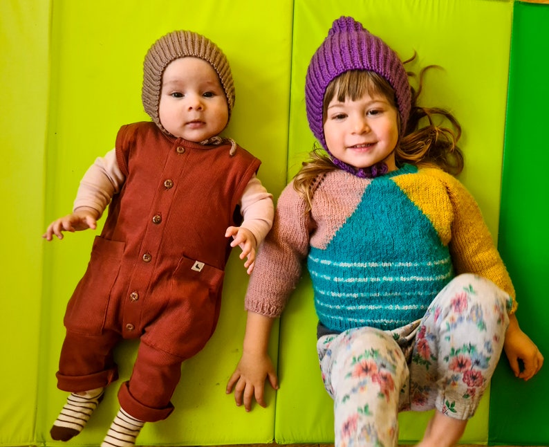 Pu&ograve; includere: Due bambini con cappelli di lana. Il bambino a sinistra indossa un cappello di lana marrone e una tuta marrone. Il bambino a destra indossa un cappello di lana viola e un maglione a righe colorate con pantaloni a fiori.