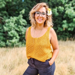 May include: A woman wearing a yellow knitted tank top with a white flower in her hair. She is standing in a field of long grass.