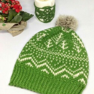 Puede incluir: Gorro de punto verde con un patr&oacute;n de copo de nieve blanco y un pomp&oacute;n de piel sint&eacute;tica marr&oacute;n.