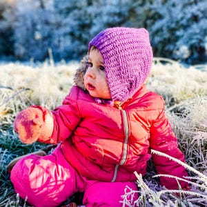 Pu&ograve; includere: Un bambino piccolo con una tuta da neve rosa e un berretto di lana viola &egrave; seduto in un campo di erba ghiacciata. Il bambino guarda a destra del fotogramma.