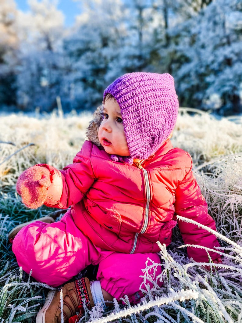 Pu&ograve; includere: Un bambino piccolo con una tuta da neve rosa e un berretto di lana viola &egrave; seduto in un campo di erba ghiacciata. Il bambino guarda a destra del fotogramma.