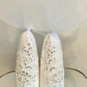 May include: Two white, cone-shaped wedding decorations with intricate lace and sequin detailing. Each cone is topped with a small bundle of tulle. The decorations are displayed on a reflective surface.