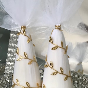 May include: Two white, cone-shaped decorations with gold leaf accents and a sheer white fabric top. The decorations are on a mirrored tray with a crystal-like border, creating an elegant display.
