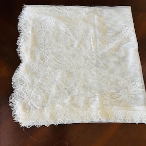 May include: Ivory lace fabric with a delicate floral pattern and scalloped edges. The lace is folded, revealing the intricate details of the design. The fabric appears to be lightweight and suitable for various craft projects or apparel.
