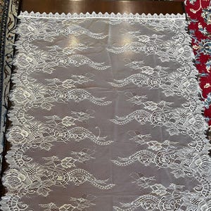 May include: Ivory lace fabric with floral and scrollwork patterns. The delicate lace features a scalloped edge and is laid flat on a dark surface. The design includes detailed flower and leaf motifs, creating an elegant textile.