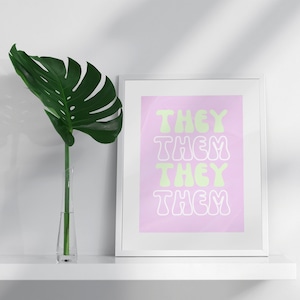 May include: A framed print with the words "THEY THEM THEY THEM" in a retro font, in light green against a pink background. A large green leaf in a clear vase is to the left of the print, on a white shelf.