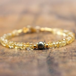 Puede incluir: Una delicada pulsera con pequeñas gemas amarillas facetadas ensartadas. La pulsera incluye una cuenta gris oscuro y detalles dorados. La joya se exhibe sobre una superficie de madera.