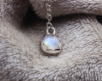 Puede incluir: Un collar de plata con un colgante cuadrado de piedra lunar iridiscente. El colgante está engastado en un bisel decorativo y cuelga de una delicada cadena. El collar se muestra sobre una superficie gris suave y texturizada.