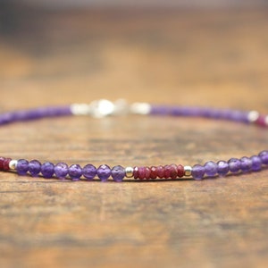 Puede incluir: Una delicada pulsera con pequeñas cuentas redondas de amatista púrpura intercaladas con pequeñas cuentas de rubí rojo y detalles plateados. La pulsera está ensartada en un cordón fino y flexible y tiene un cierre plateado.