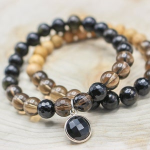 May include: Two beaded bracelets. One bracelet features dark brown, faceted beads and a silver charm with a black, faceted stone. The other bracelet has light brown, round beads. Both bracelets are on a light wood surface.