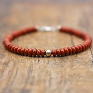Peut inclure: Un bracelet en perles de jaspe rouge avec un fermoir argenté et une seule perle argentée. Le bracelet est composé de petites perles rondes brun-rouge. Le fermoir est argenté et la perle unique est centrée.