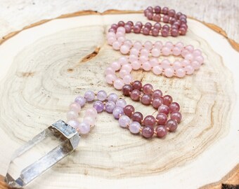 May include: A collection of gemstone bead necklaces in shades of pink, purple, and clear, displayed on a light-colored wooden surface. The necklaces feature various sizes of round beads and a clear crystal pendant.
