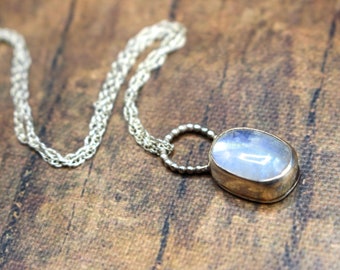 Puede incluir: Un collar de plata con un colgante de piedra lunar. El colgante ovalado tiene un tono azul claro. La cadena es de diseño trenzado. El collar se muestra sobre una superficie de madera marrón.