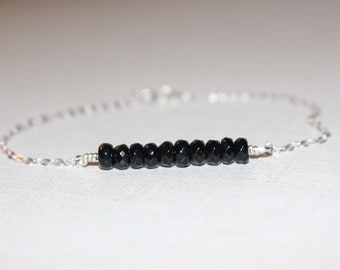 May include: A delicate bracelet featuring a series of small, faceted black beads strung together. The beads are centered on a fine, silver-colored chain. The bracelet is designed to be worn around the wrist.
